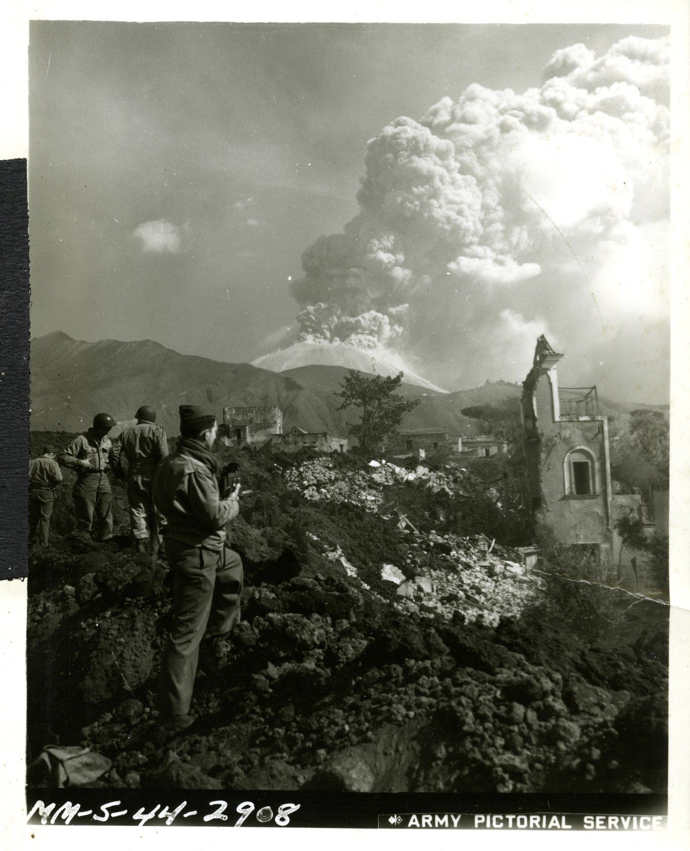 Soldati americani guardano le macerie di San Sebastiano al Vesuvio coperte di lava, mentre sullo sfondo il vulcano emette una grande nube